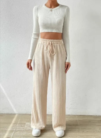 Cream wide-leg drawstring pants with vertical texture, worn with a cropped cream long-sleeve top and white sneakers, standing against a textured white wall on a light wood floor.